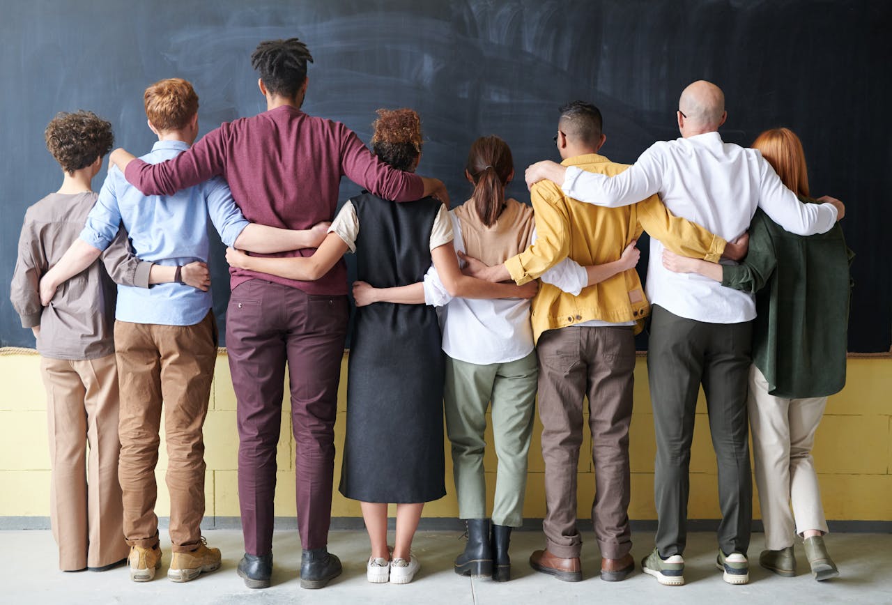 gallery-4 A diverse group of adults in casual outfits hugging in front of a chalkboard, symbolizing teamwork.