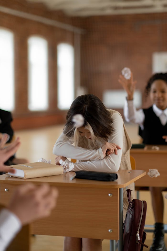 why-choose-us A teenage girl sits at her desk, visibly upset as classmates throw paper balls at her.